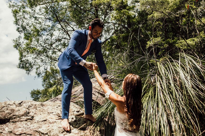 Groom helps bride onto rock outlook in rainforest